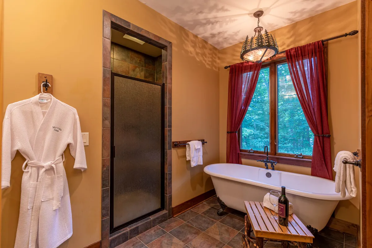 A spa-like bathroom featuring a free-standing bathtub positioned next to a large window with a scenic view and illuminated by a statement chandelier. The room is flooded with natural light, evoking both luxury and tranquility—an ideal place for a soothing soak at day's end.
