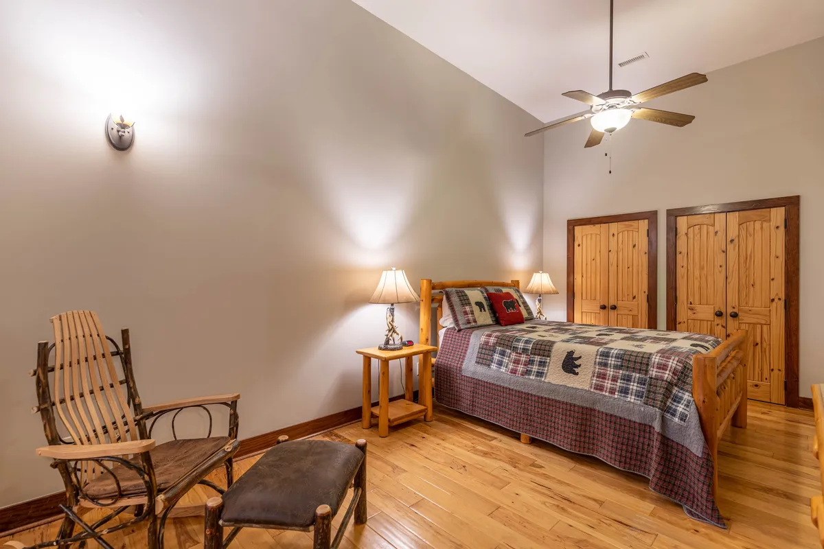 A welcoming bedroom scene with a bed draped in a cozy plaid blanket, flanked by wooden bedside tables with warm lighting, a classic wooden rocking chair, and rustic closet doors. The atmosphere is inviting and restful, perfect for relaxing or reading after a long day.