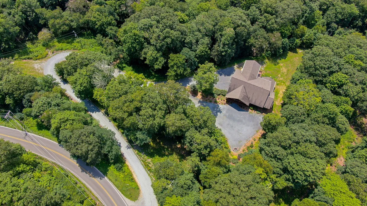 A tranquil exterior view showing the house nestled among tall trees, with a curving road leading through the lush greenery. The scene hints at peaceful seclusion while also suggesting easy road access to nearby attractions.