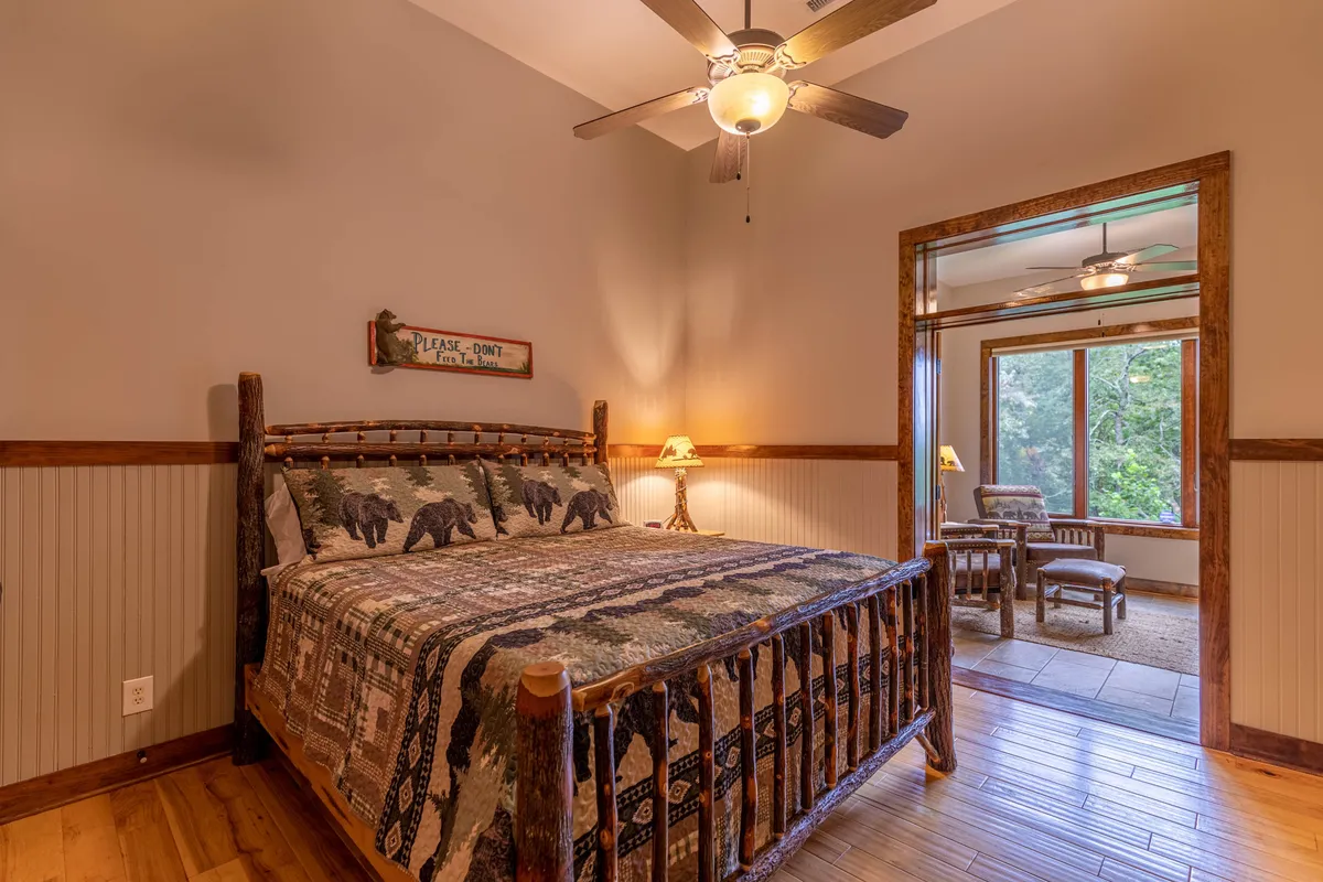 A rustic bedroom with a wooden bed adorned in a bear-themed quilt, a nightstand with a glowing lamp, and a window offering pastoral views. The scene is cozy and serene, ideal for families or guests seeking a tranquil night's rest.