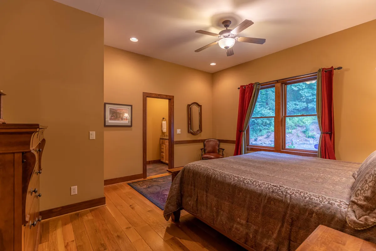 A light-filled bedroom with a plush bed positioned by a large window, an inviting chair, and tasteful décor including a decorative mirror. The atmosphere is peaceful and perfect for restful mornings or winding down in privacy.