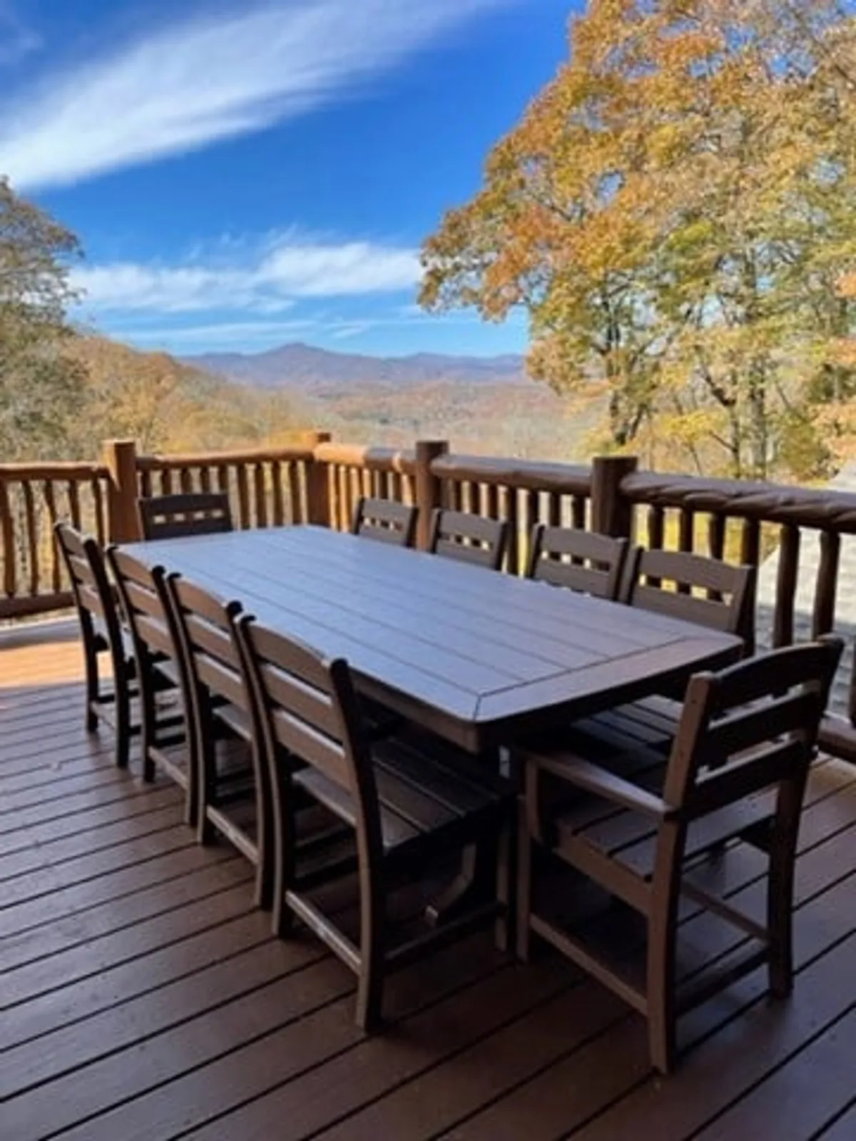 An expansive wooden dining table surrounded by ten seats, set outdoors against a dramatic mountain and autumn foliage backdrop. The setting is perfect for communal meals, with enough space for large gatherings and alfresco dining.