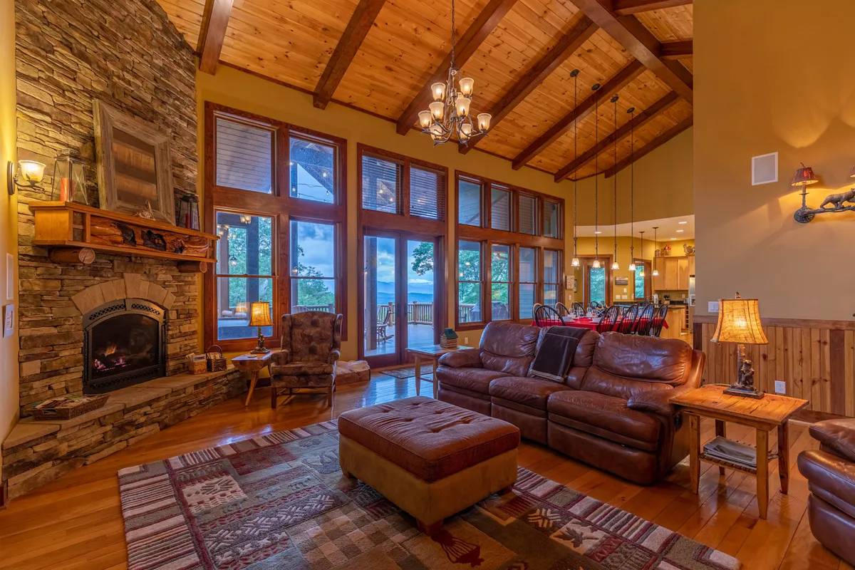 The image showcases a spacious living room with a dramatic stone fireplace as a central feature. Large windows bring in natural light and frame the mountain scenery. Leather couches and a handcrafted coffee table create a cozy area for group gatherings, while exposed wooden beams and rich textures add rustic elegance to the space.