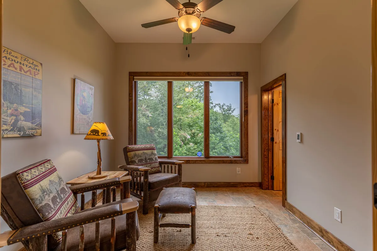 A sun-drenched sunroom with large windows revealing a serene view of lush trees outside. Comfortable wooden chairs and a rustic lamp invite relaxation, while the space is filled with natural light and a calming ambiance perfect for unwinding or enjoying nature's tranquility from indoors.