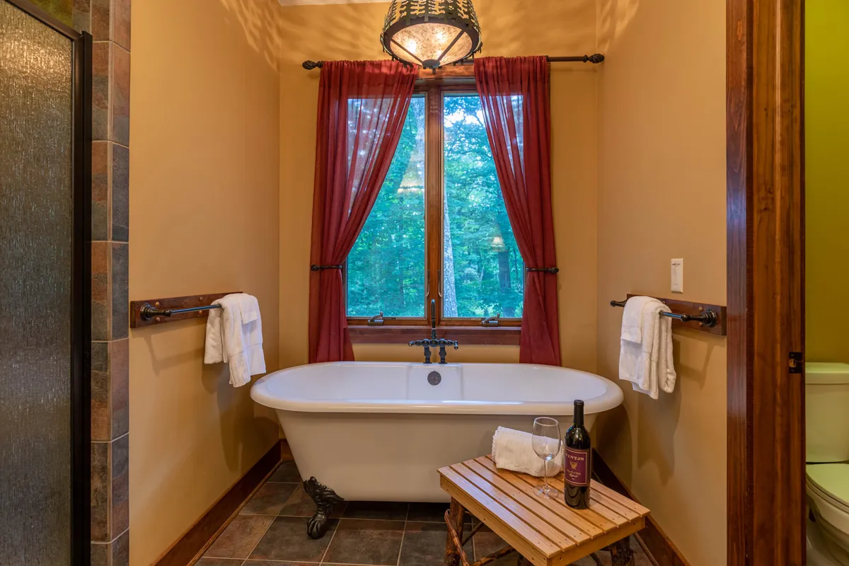 A serene bathroom scene featuring a freestanding claw-foot bathtub next to a large window, with a wooden stool holding a glass of wine and a book. The setting bathes in soft light, creating a luxurious, spa-like atmosphere for relaxation.