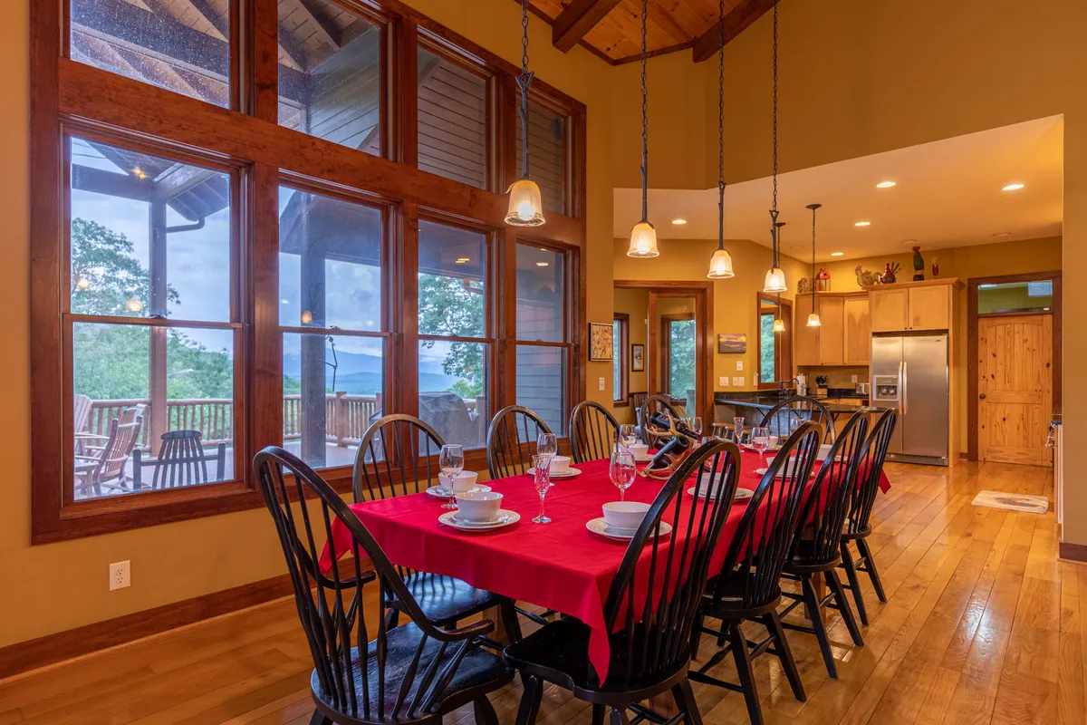 The open-concept dining and kitchen area features a large dining table suitable for ten, floor-to-ceiling windows overlooking the mountains, and a warm wooden ceiling. The adjacent kitchen area is visible, emphasizing the seamless flow between cooking, dining, and social spaces, ideal for celebrations or family meals.