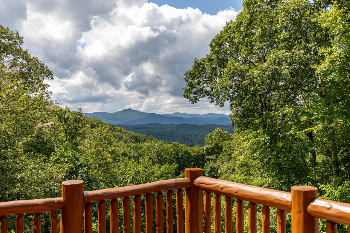 A peaceful deck scene with a wooden railing in the foreground, lush green trees, and a panoramic mountain range stretching across the horizon. The scene evokes the natural beauty and scenic vistas of the Blowing Rock area.