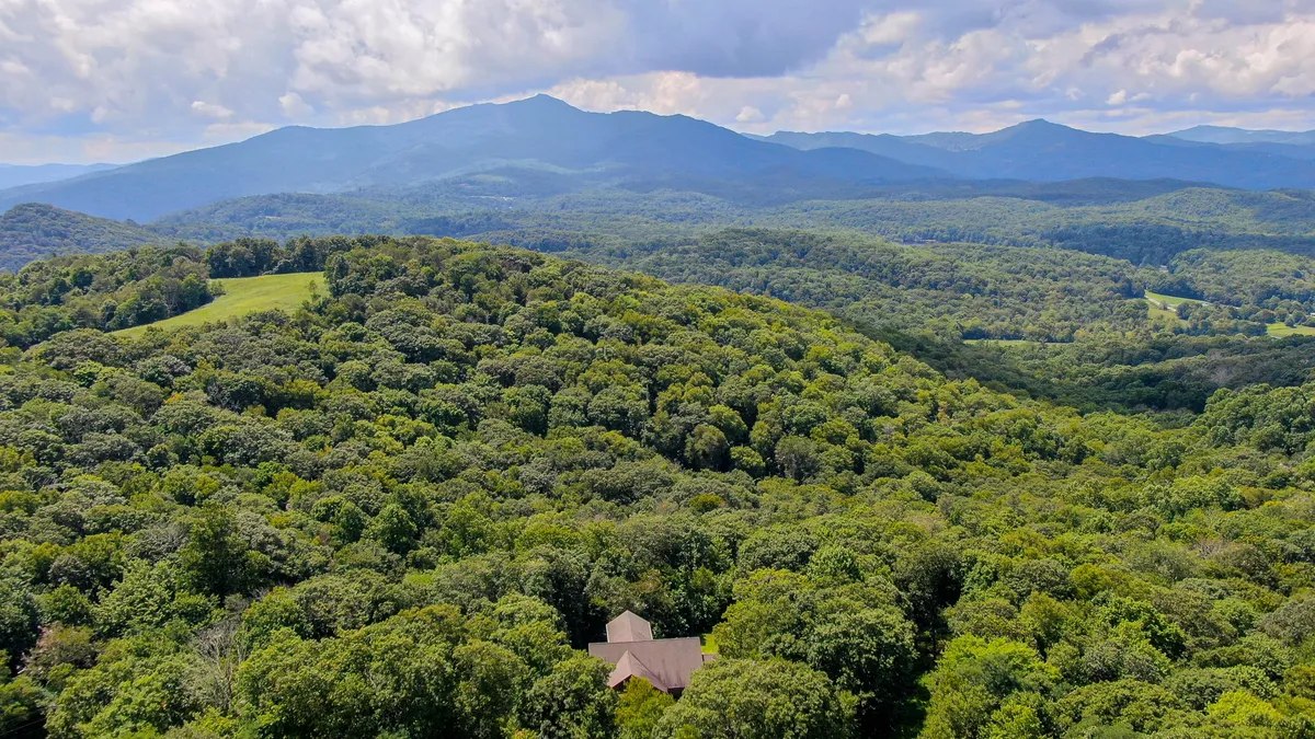 A sweeping view of rolling hills blanketed with dense forest, distant blue mountains on the horizon, and a sky scattered with clouds. The natural landscape evokes the wild, untouched beauty that surrounds the property and hints at the hiking adventures possible directly from the site.