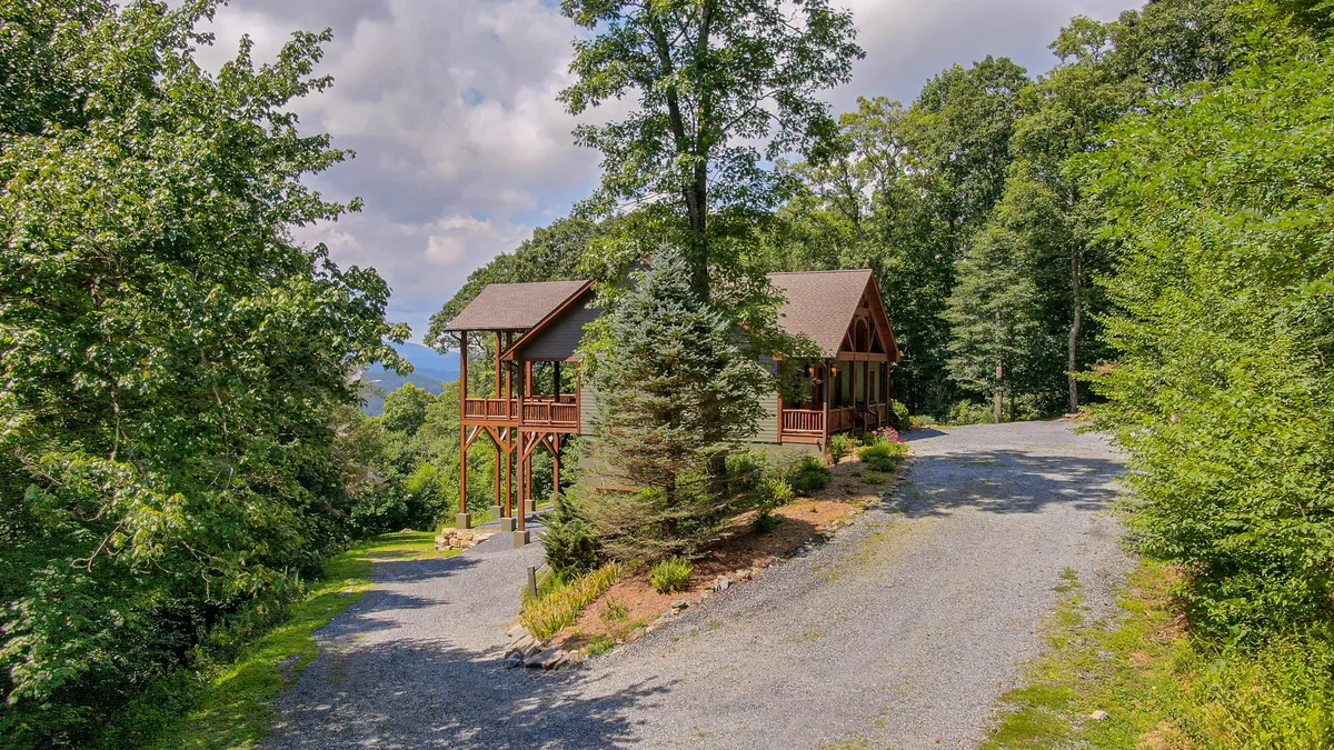 A broad exterior view of the wooden house embraced by mature trees, with a visible pathway leading into the woods. The image captures the connection to nature and hints at the proximity of hiking trails.
