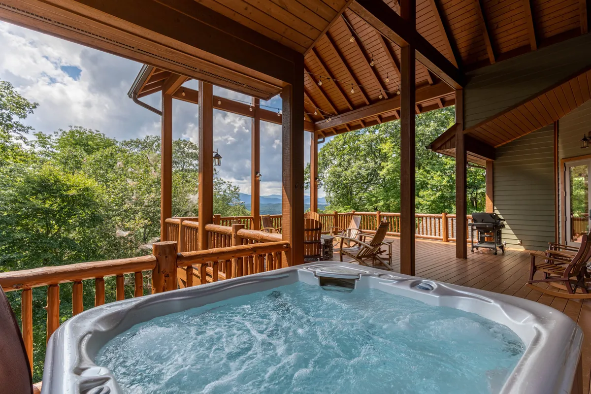A spacious wooden deck featuring a five-person hot tub, comfortable outdoor seating, a grill, and sweeping views of trees and distant blue mountains. The arrangement invites relaxation and socializing, with natural wood finishes blending seamlessly into the forested backdrop.