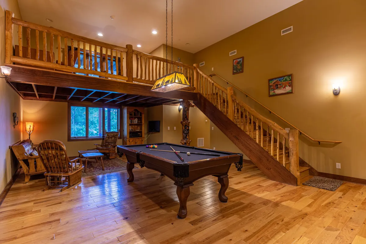This image showcases a spacious game room featuring a pool table, wooden staircase, a cozy seating area, and decorative lighting. The setting is designed for socializing, friendly competition, and relaxed gatherings, with a welcoming and lively atmosphere.